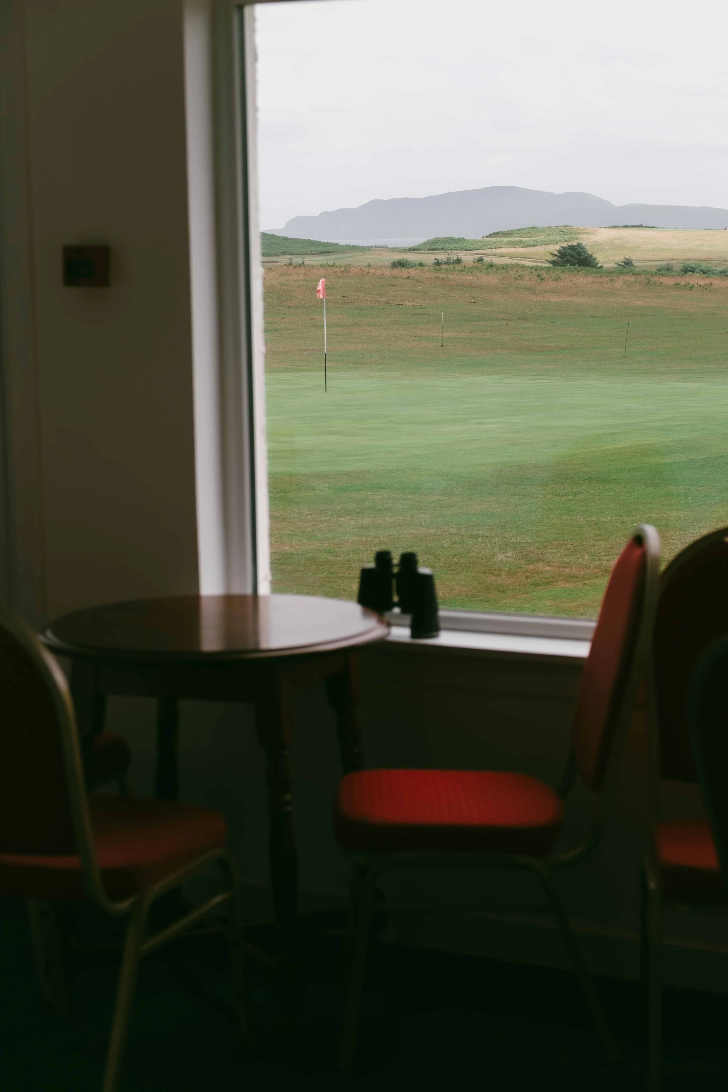 Interior of a building with a round table and four red chairs, looking out a window at a golf course with green grass, flags, and mountains in the background.