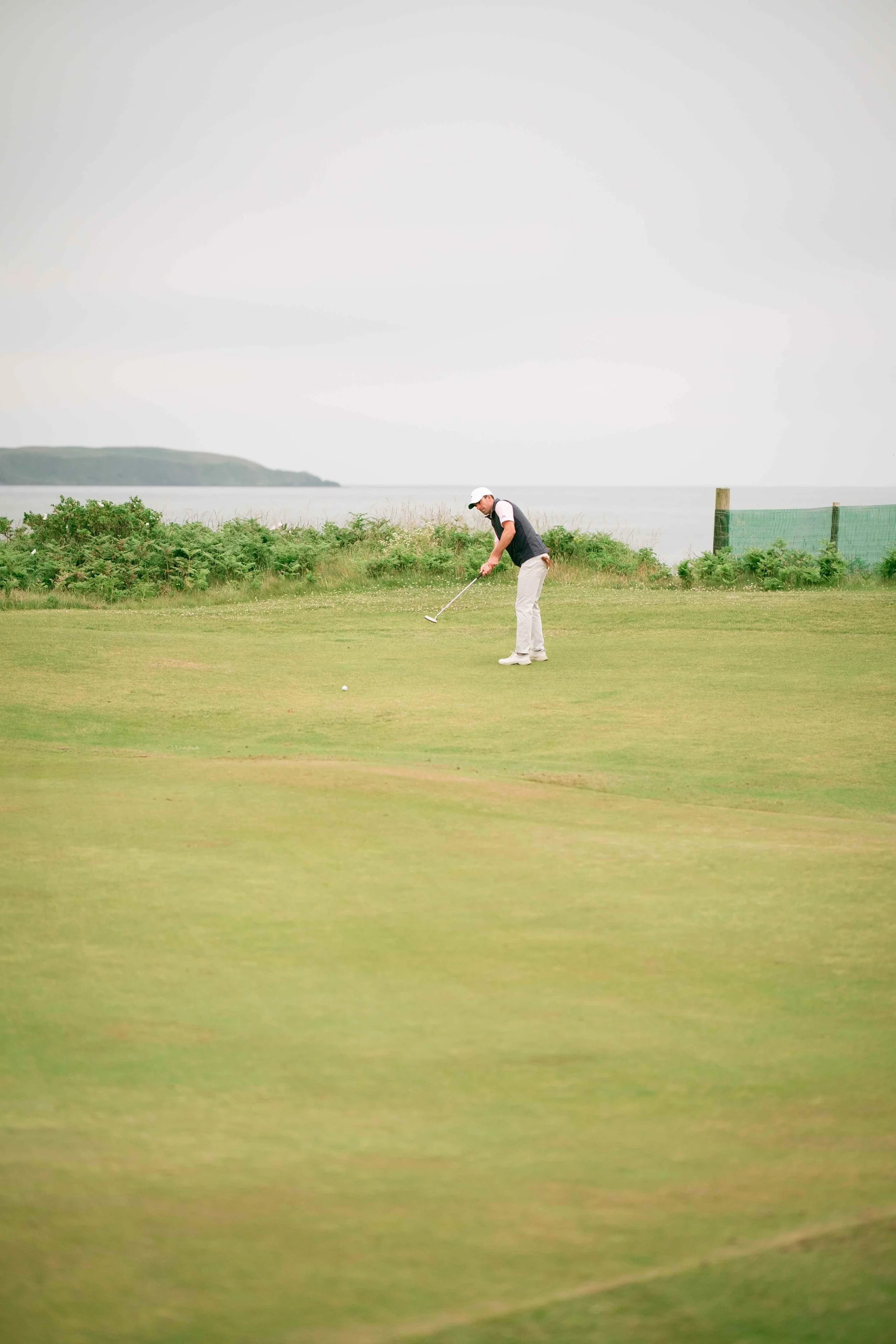 A man playing golf on a grassy course near water with an overcast sky.