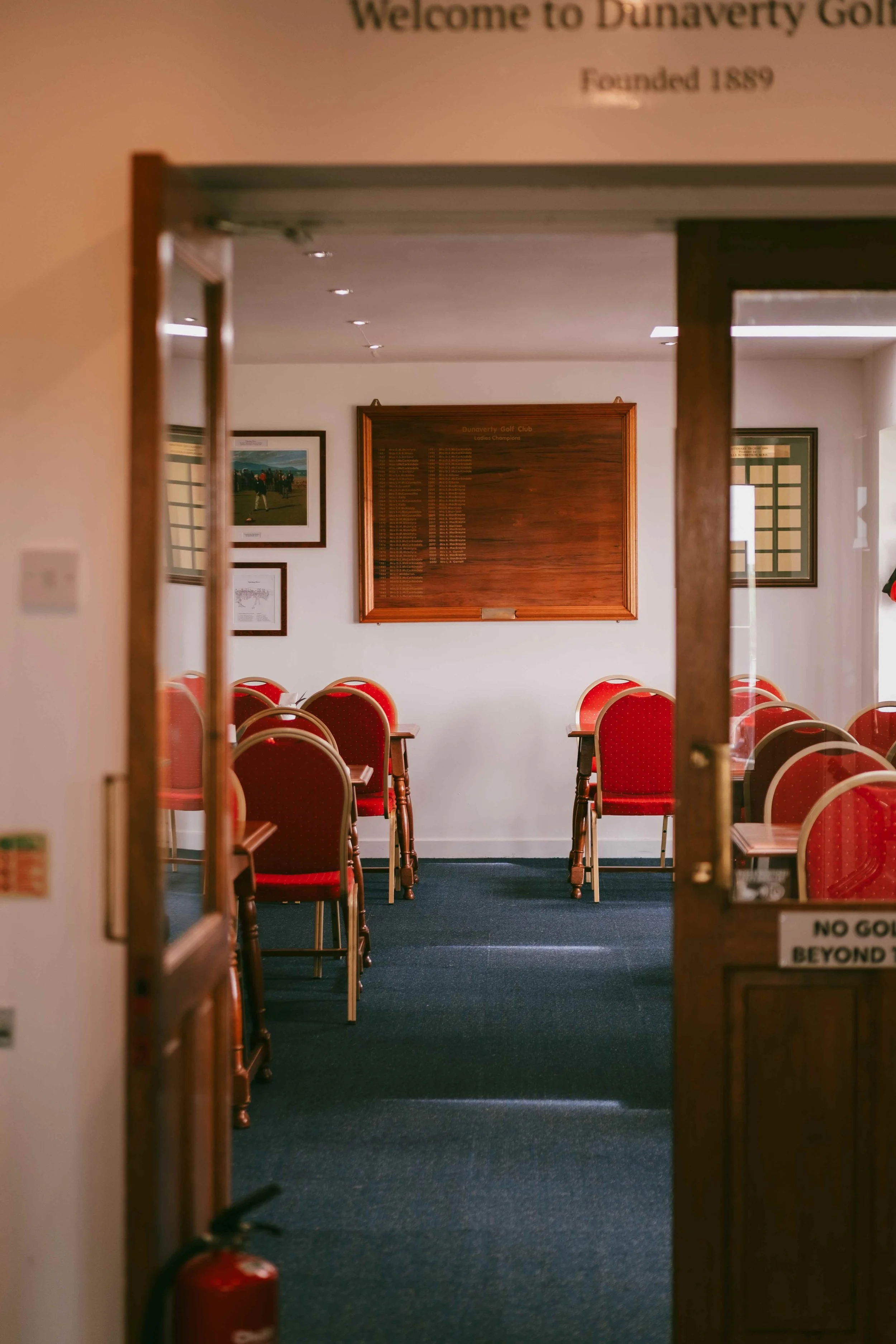 A view through a doorway into a room with rows of red cushioned chairs and a wooden plaque on the wall displaying club championship results at Dunavery Golf Club. The room has framed pictures on the walls, and natural light is coming from the right.