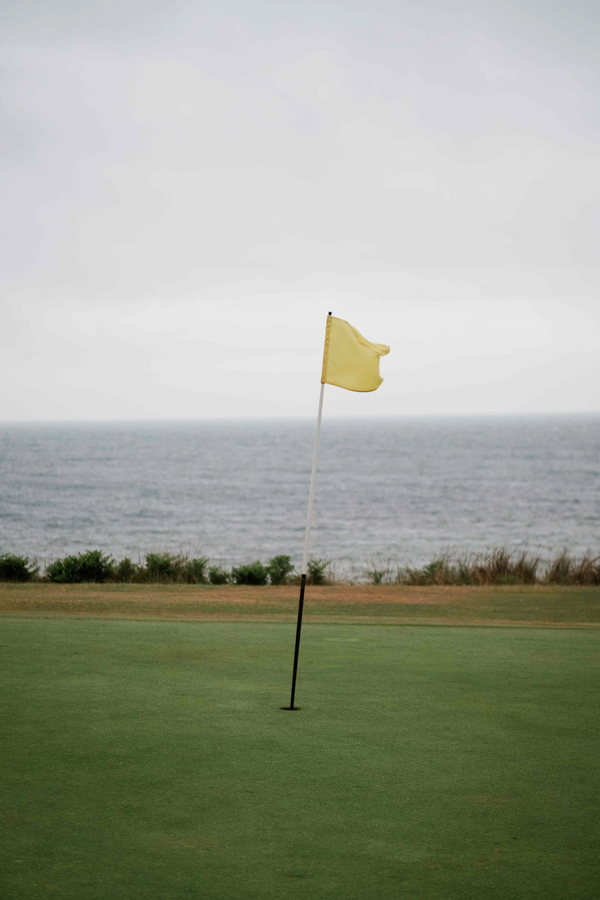 A yellow golf flag on a putting green near the ocean with clouds in the sky.