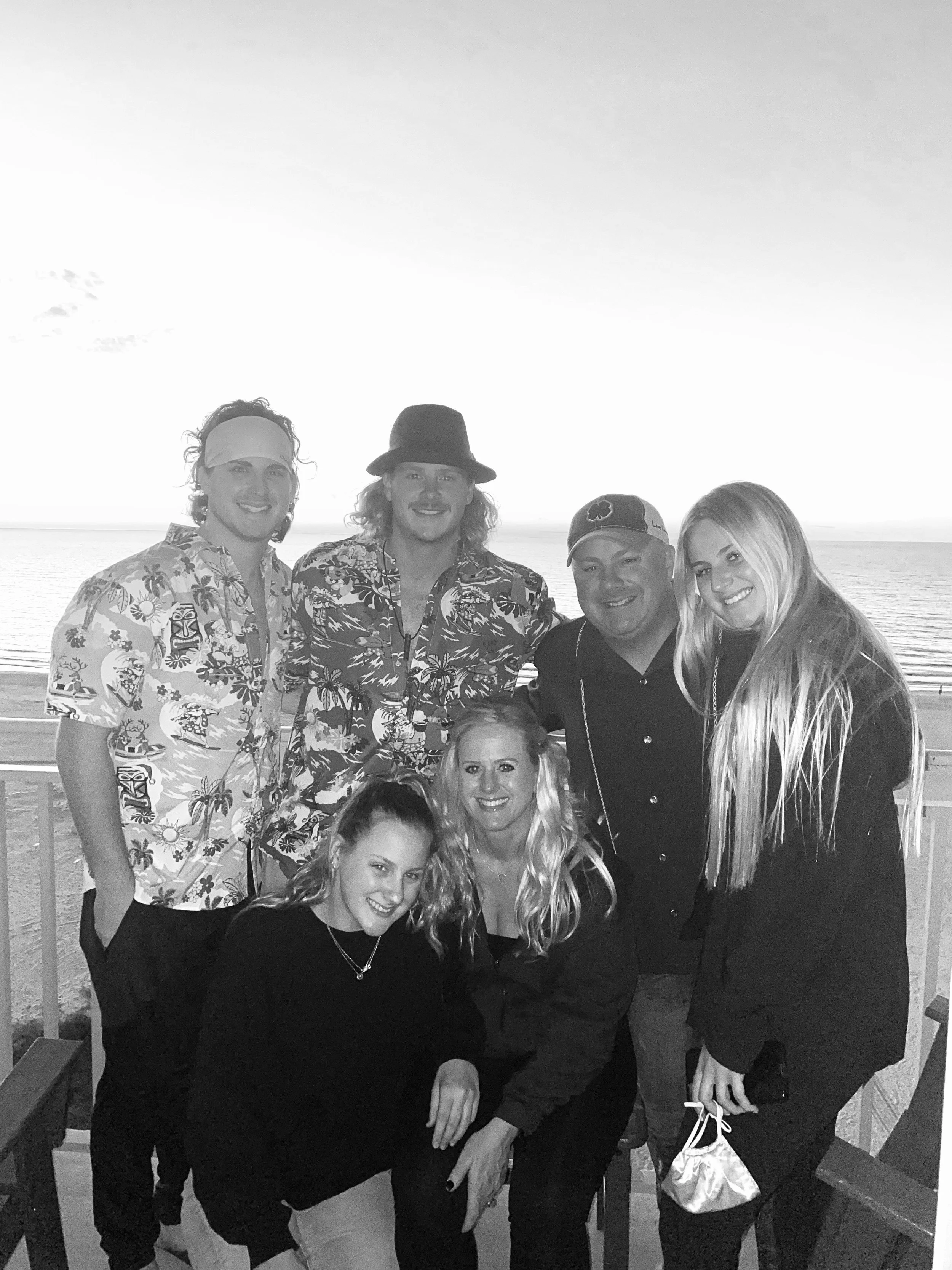 Group of eight people standing on a beach at sunset, smiling for the photo, dressed in tropical clothing.