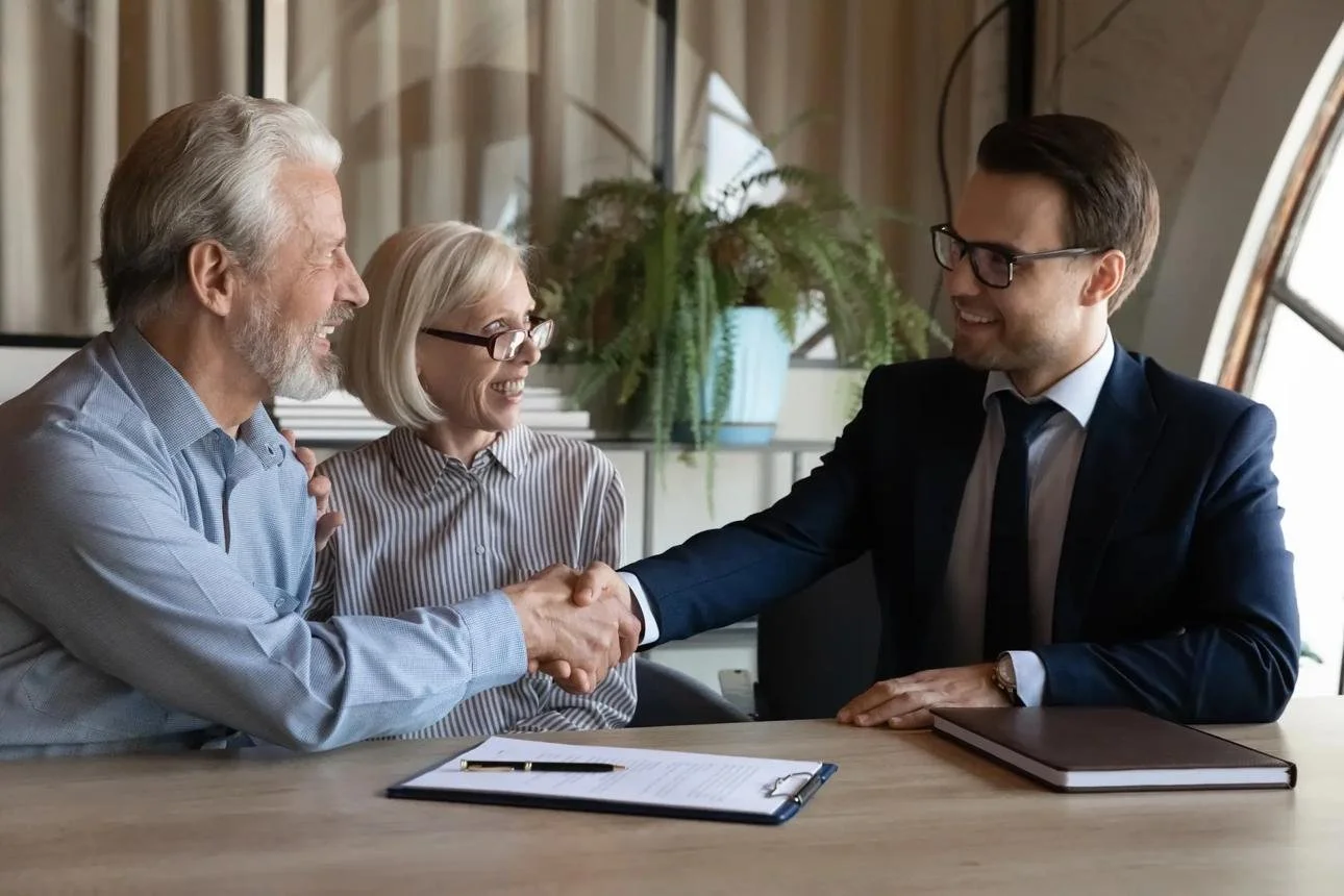 Older man and woman shaking hands with a younger man in a business meeting. They are smiling and sitting at a table with documents and a notebook.