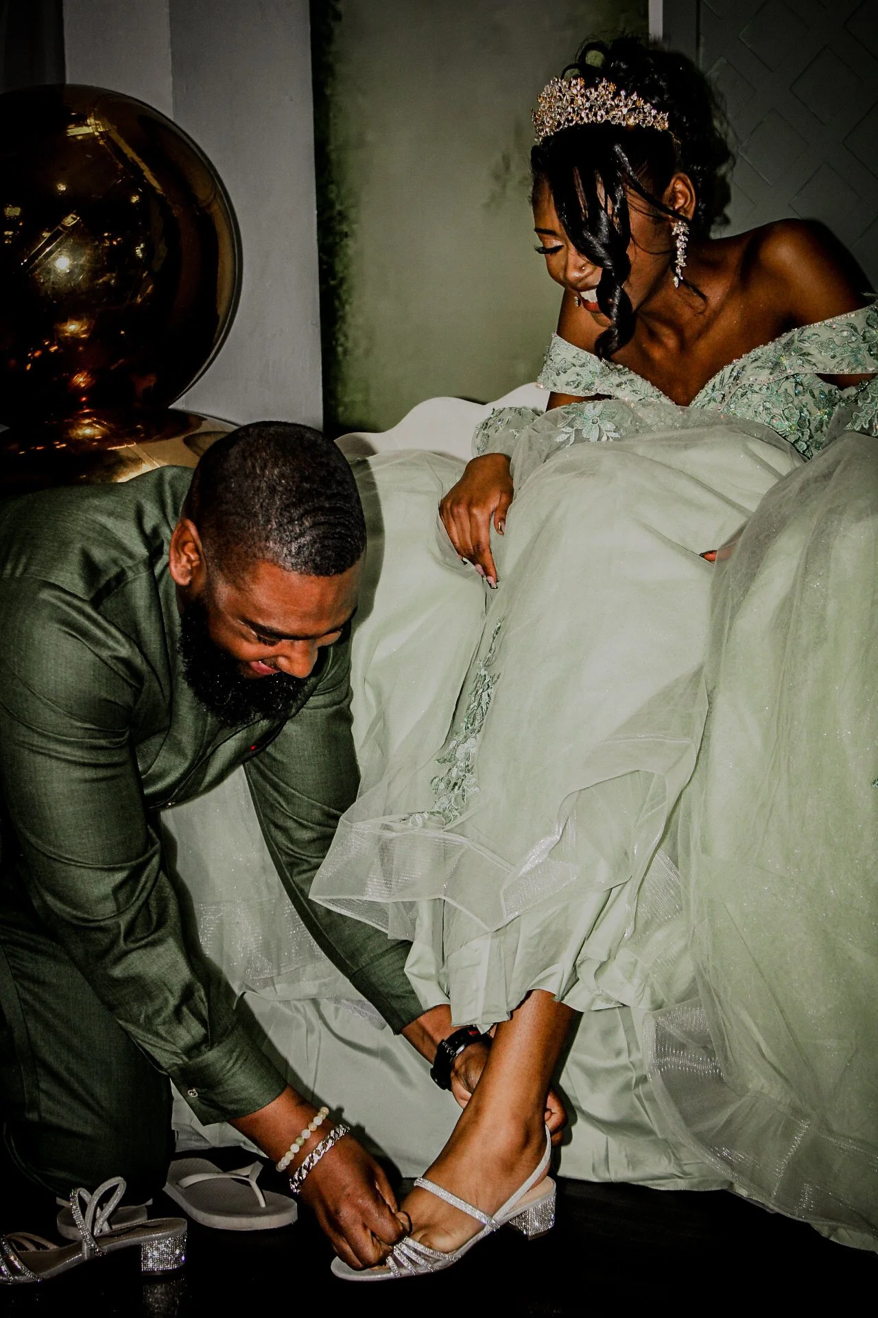 A woman in a white wedding dress being assisted by a man in a dark green suit as she puts on her silver high-heeled shoes. She is wearing a tiara and jewelry, with her veil lifted. The man is kneeling, helping her with her shoe, in an indoor setting.