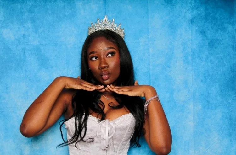 A woman in a white dress wearing a crown making a playful kissy face against a blue background.