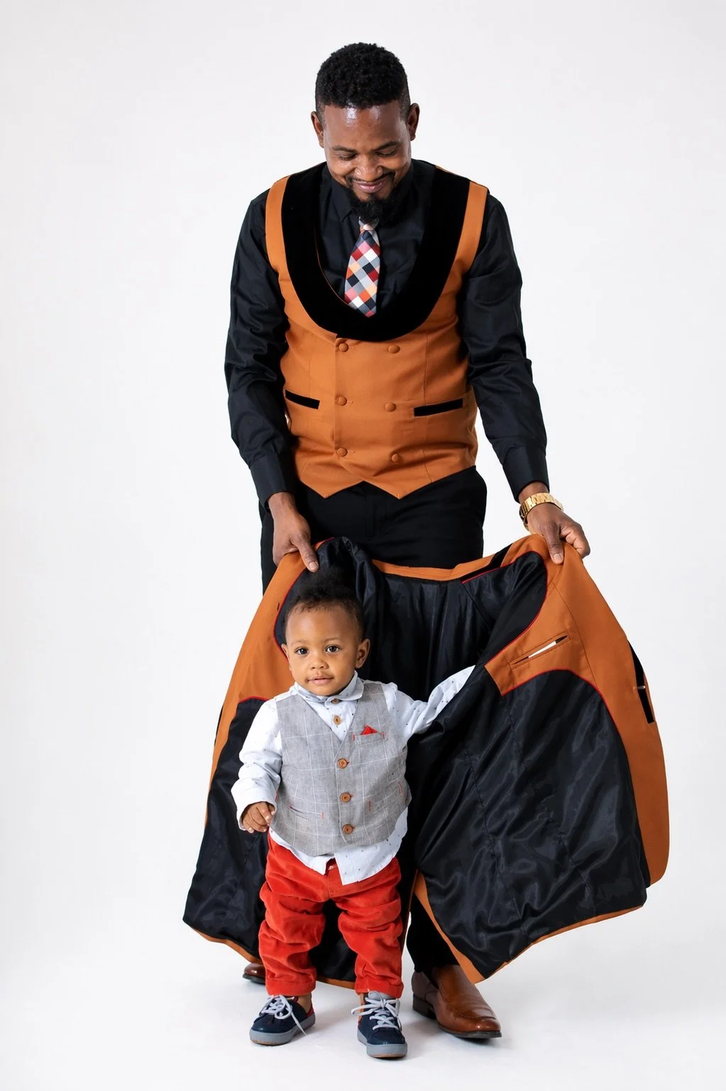 A man in a stylish orange and black vest and black shirt is smiling as he reveals a surprise outfit for a young boy, who is standing inside a large orange and black bag, wearing a grey vest, white shirt, red pants, and sneakers.