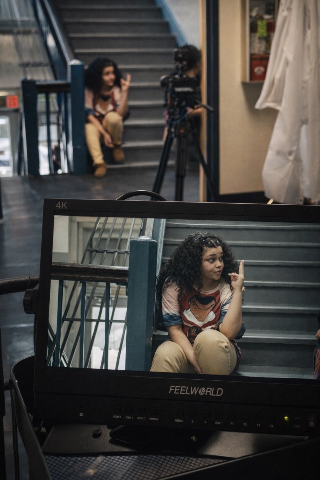 A young girl with curly dark hair sitting on stairs, wearing a Wonder Woman t-shirt, and showing a finger gesture while looking to the side, with a video monitor in the foreground reflecting her image, and a person with curly hair and camera in the background.