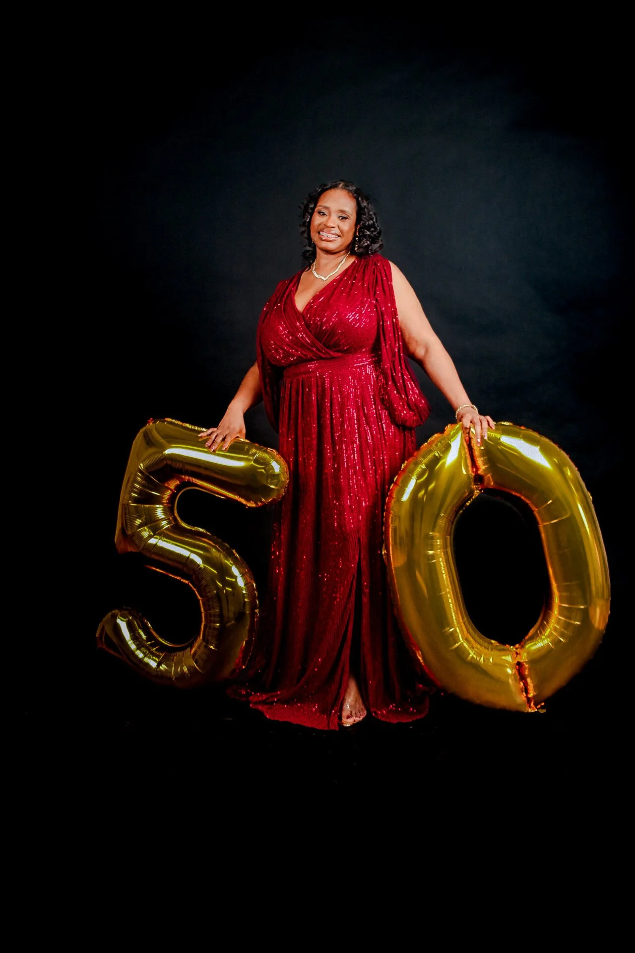 A woman in a red, sequined gown holding gold number balloons '5' and '0' against a black background.