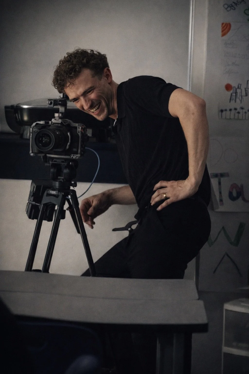A man with curly hair and a big smile, wearing a black shirt, is adjusting a camera on a tripod in a dark room with a whiteboard in the background.