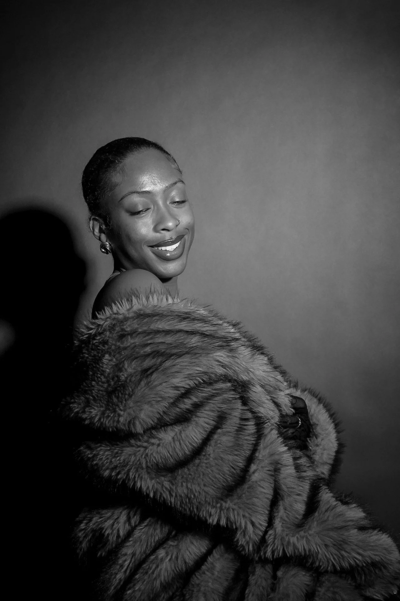 Black and white portrait of a woman smiling with closed eyes, wearing earrings and a fur coat, against a plain background.