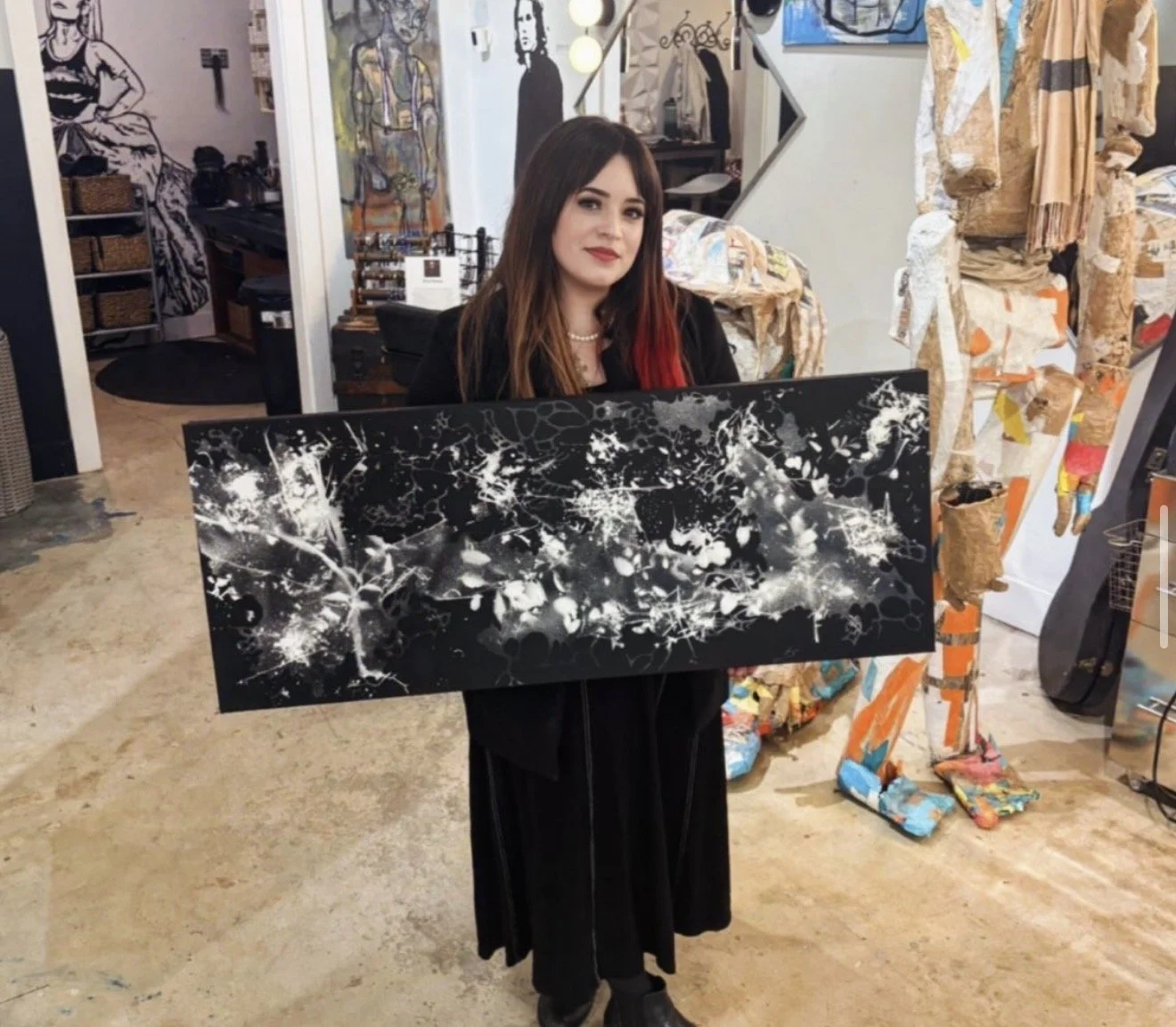 Woman with brown and red hair holding abstract black and white painting in art studio with colorful sculptures and paintings in the background.