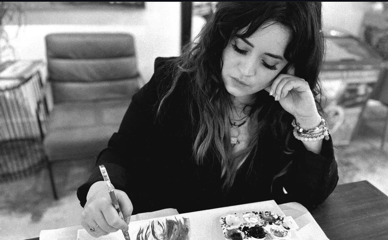 A woman with wavy hair and bracelets, sitting at a table, drawing or painting with a brush on paper with paint splotches.