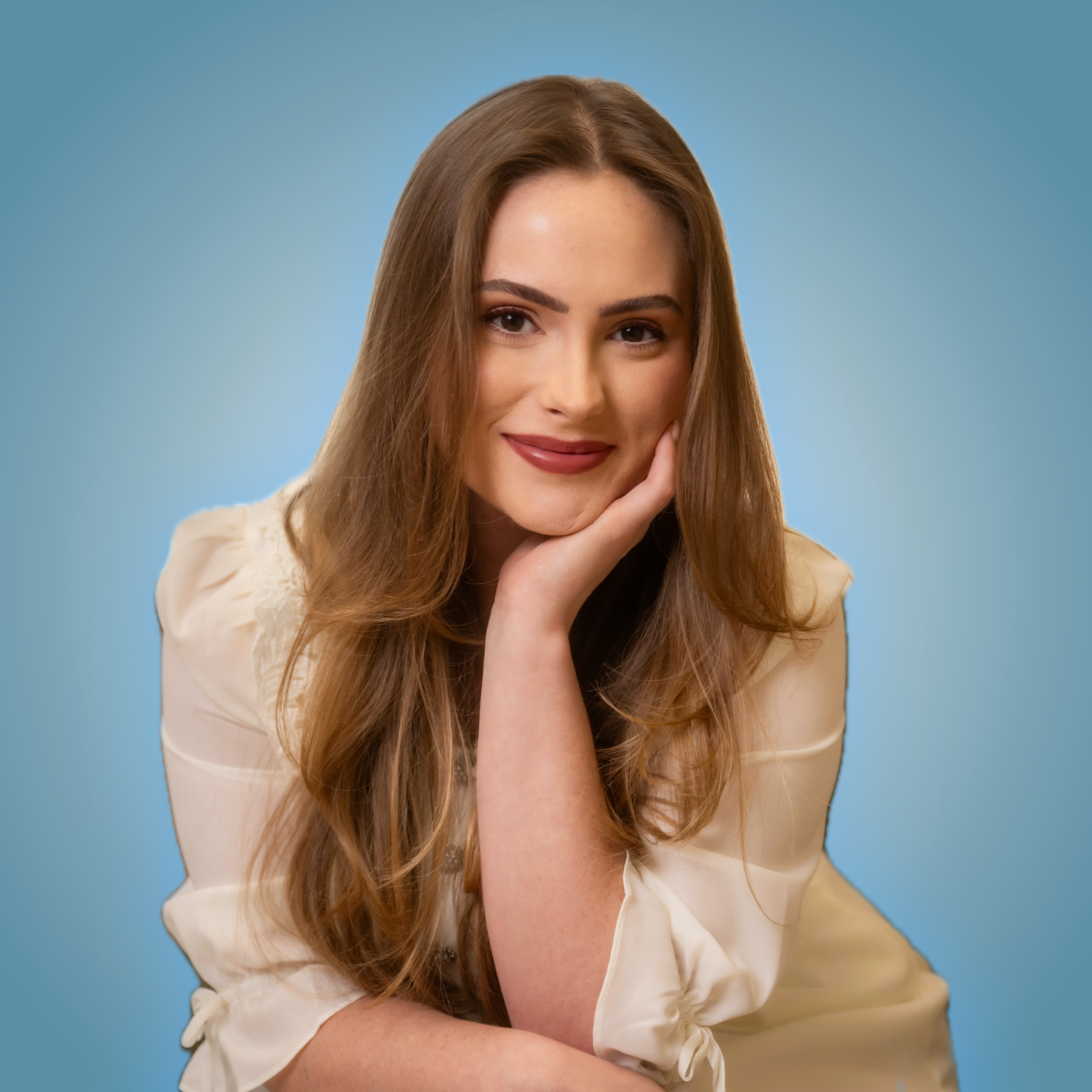 Portrait of a young woman with long brown hair, smiling softly with her hand gently touching her face, wearing a light-colored top, against a blue background.