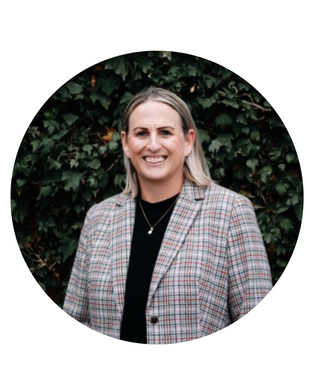 A woman with shoulder-length blonde hair, smiling, wearing a plaid blazer over a black top, standing outdoors in front of green leafy bushes.