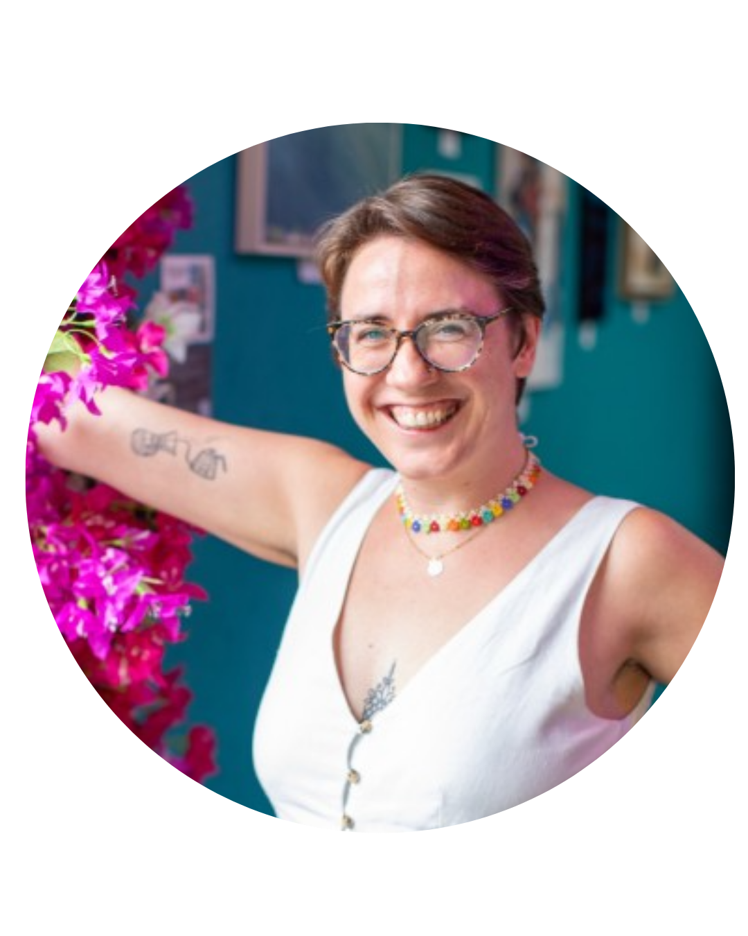 Smiling woman with short brown hair, glasses, colorful beaded necklace, white sleeveless top, tattoos on arm and chest, beside pink flowers in an indoor setting.