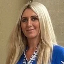 A woman with long blonde hair wearing a blue top standing against a beige wall.