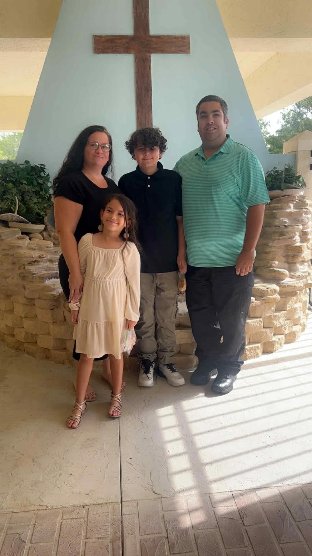 A family of four standing inside a church with a cross on the wall behind them. The family includes a woman, a girl, a boy, and a man. The woman and the girl are smiling, and the boy and the man are standing close together.