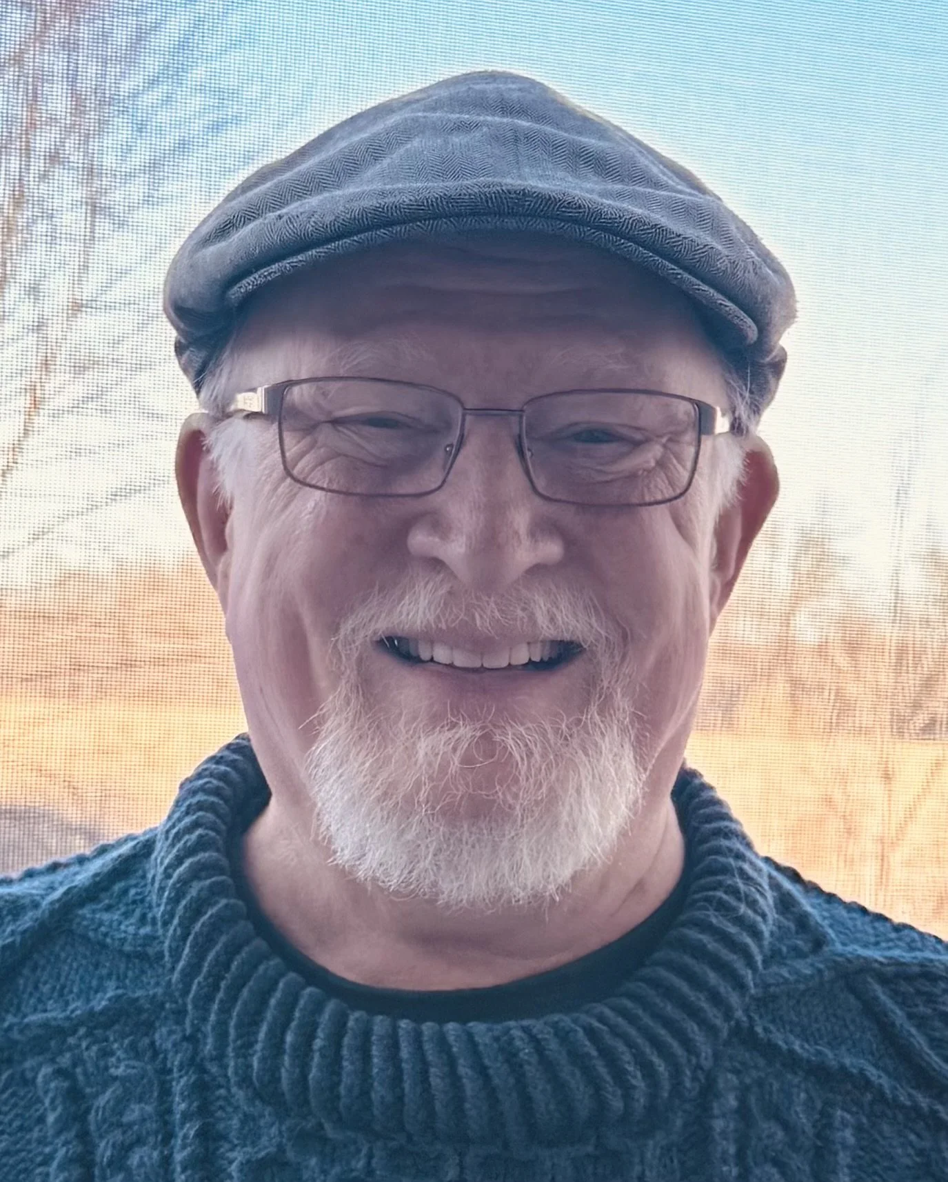 An older man with glasses, a gray beard, and a cap smiling outdoors during sunset.