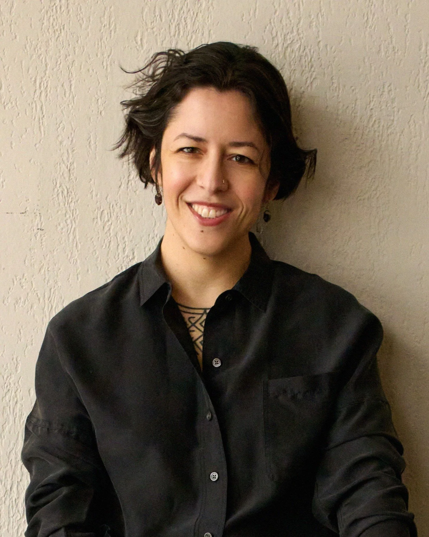 A woman with short dark curly hair and earrings smiles at the camera, wearing a black shirt, standing against a textured beige wall.