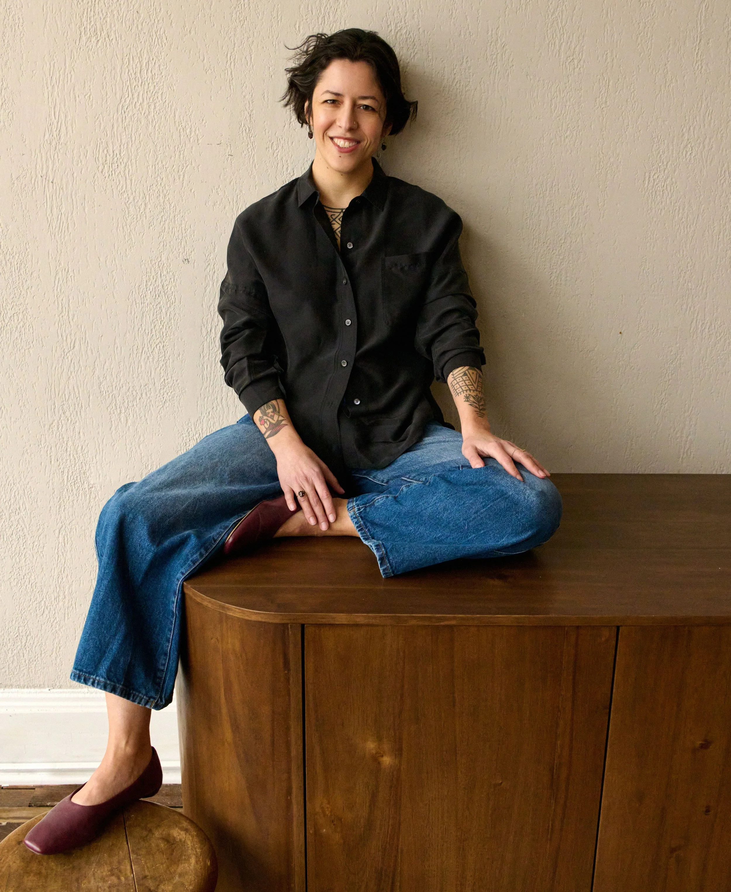 A woman with short dark hair, tattoos on her arms, and wearing a black shirt and blue jeans, is sitting cross-legged on a wooden table against a beige textured wall. She is smiling and winking at the camera.