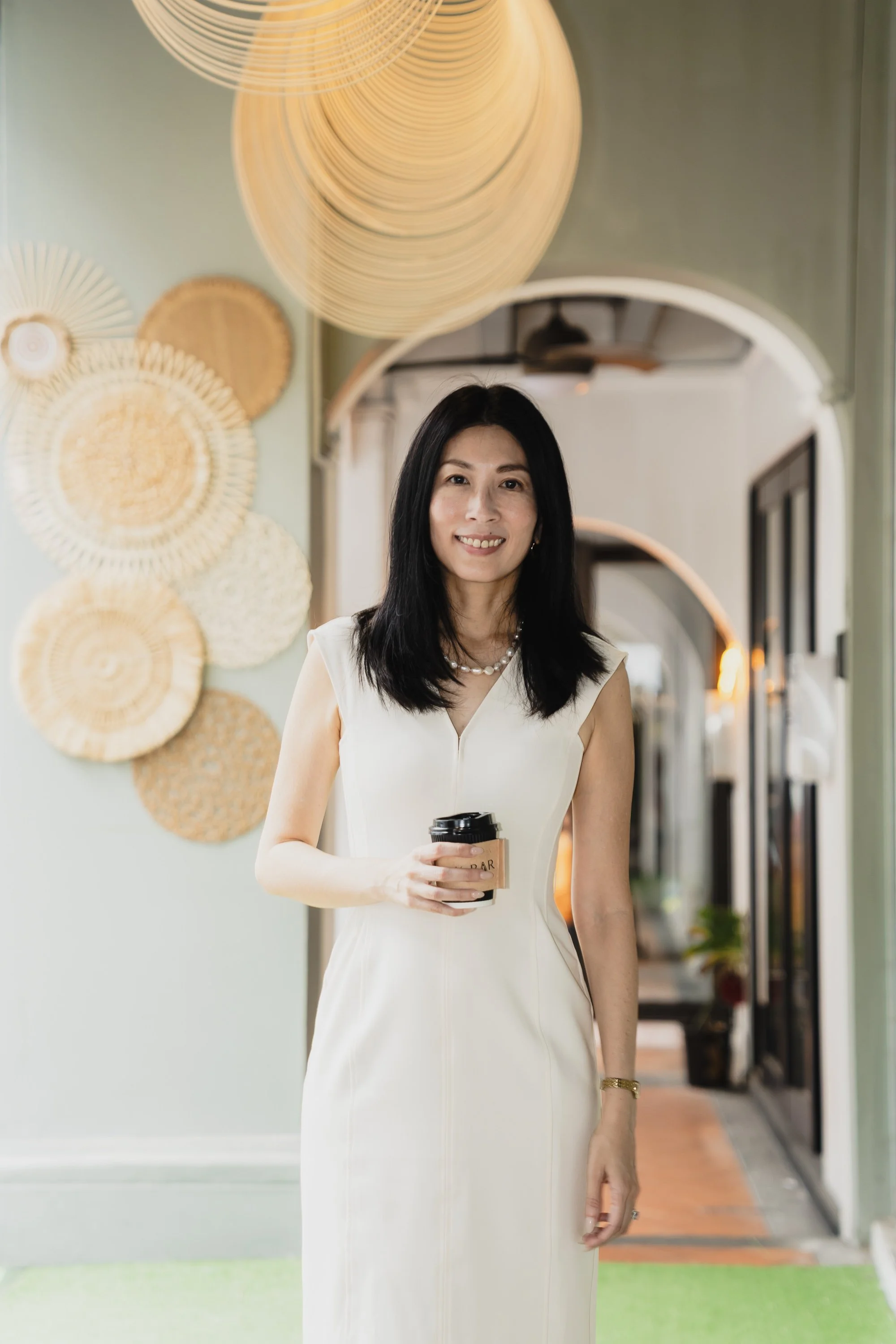 A woman with long black hair smiling while holding a coffee cup inside a stylish cafe or restaurant. The background features decorative circular wall art and an arched doorway.