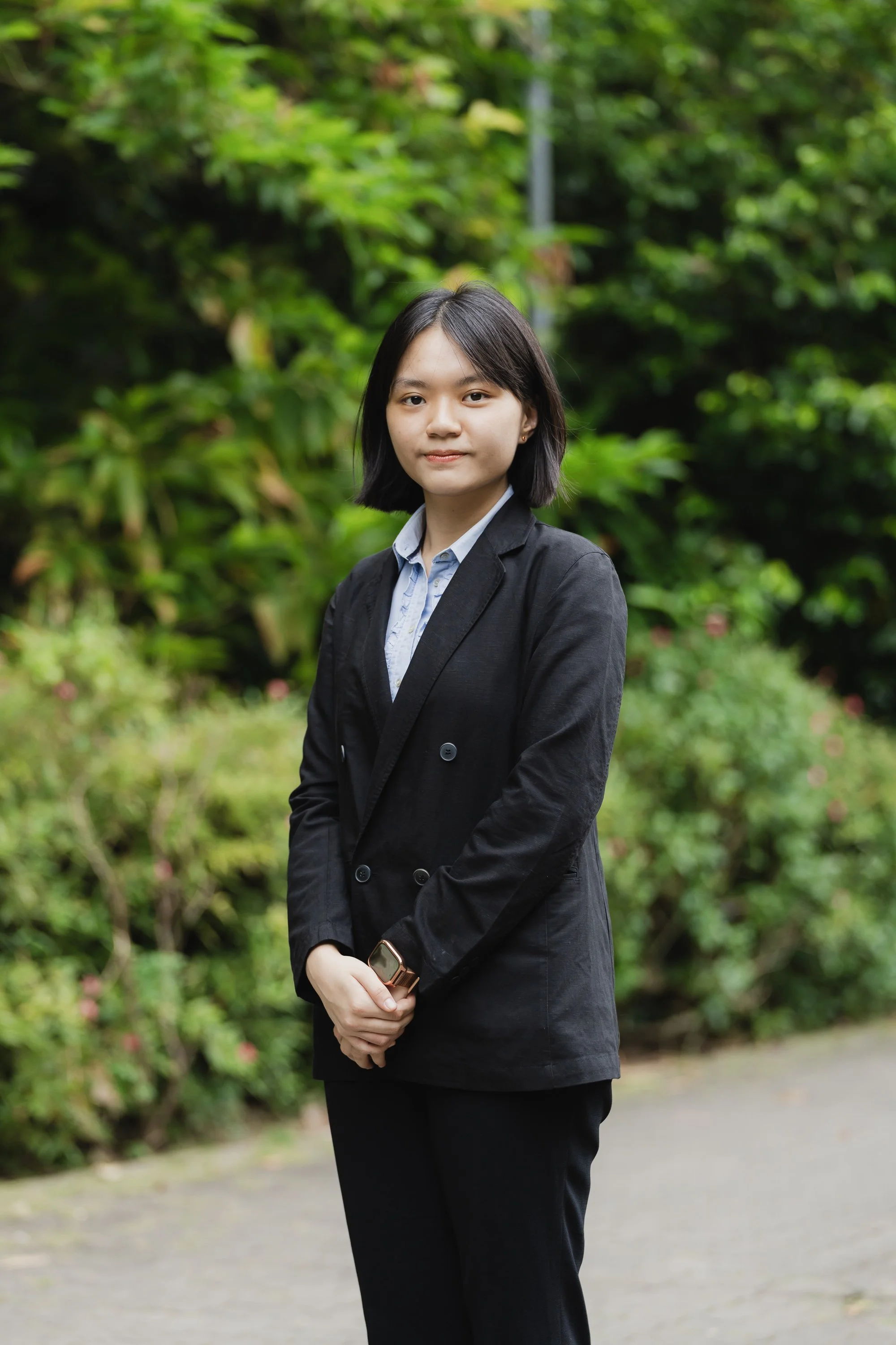 A young woman with shoulder-length black hair and light skin, wearing a black blazer and a light blue shirt, standing outdoors in front of a lush green background with trees and bushes.