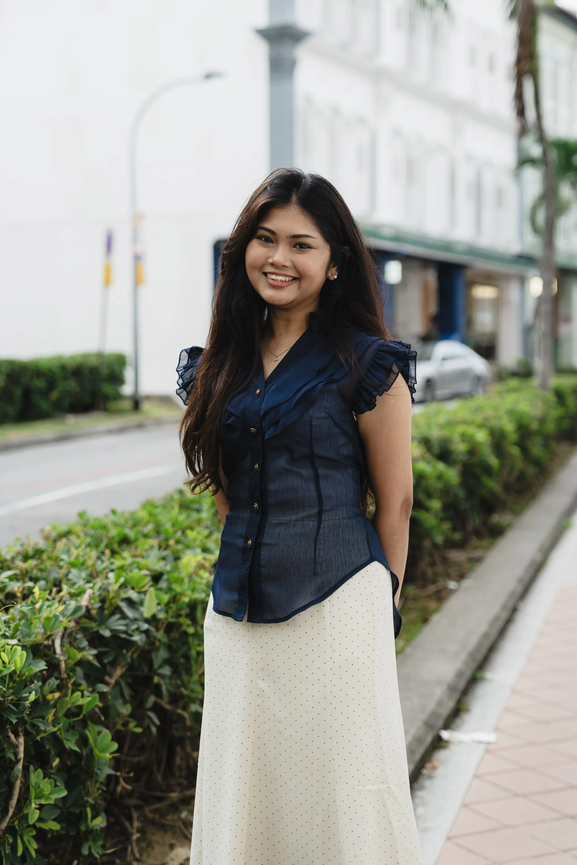A woman standing outdoors near a curb with greenery, smiling, wearing a navy blue top and white dotted skirt.