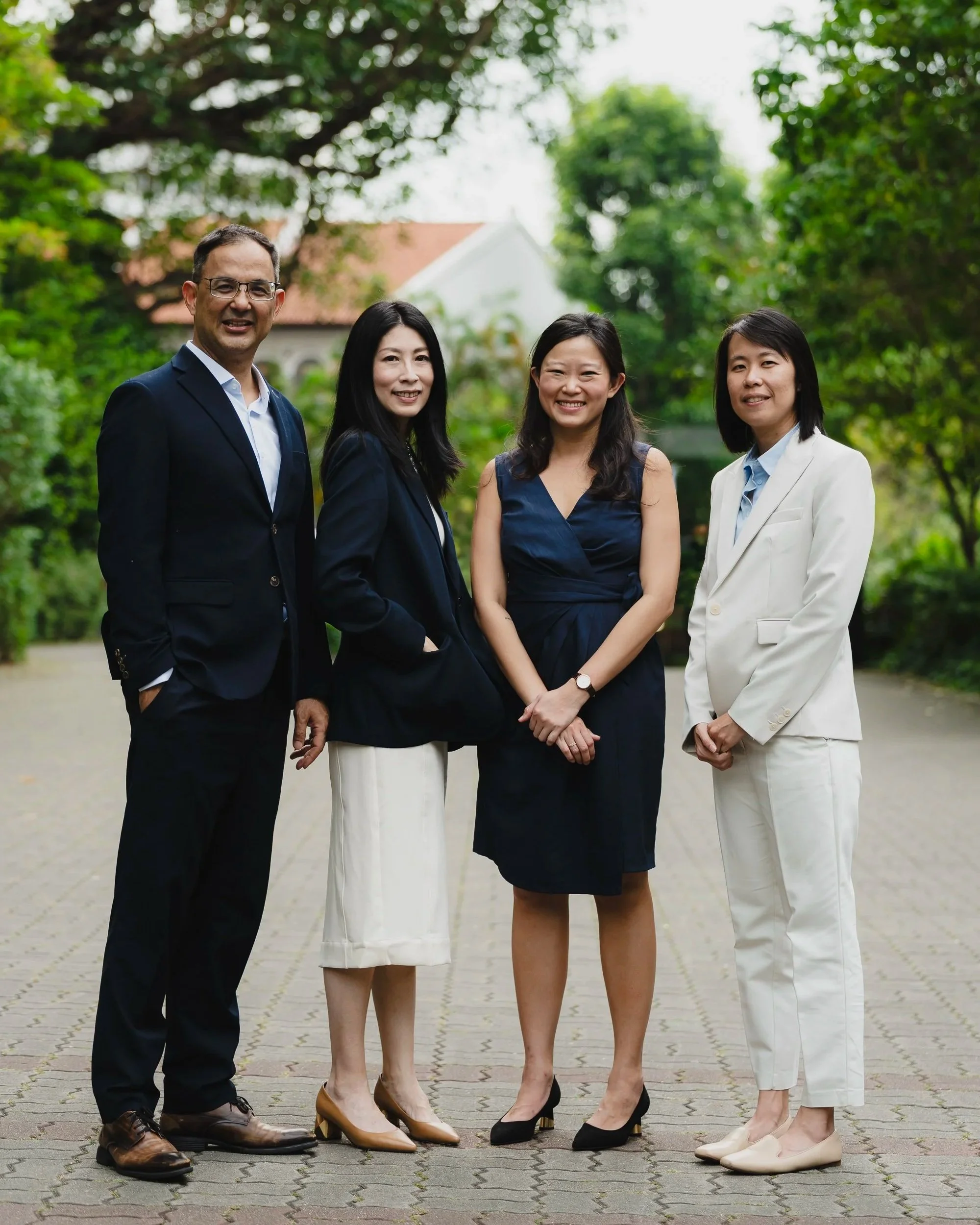 Four professionally dressed individuals, two men and two women, standing outdoors on a paved path with trees and a building in the background. They are smiling and facing the camera.