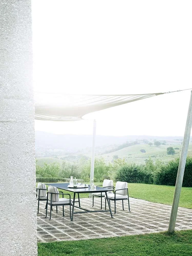 Outdoor patio with a black table, four white chairs, and a large white shade sail, overlooking green rolling hills and trees.