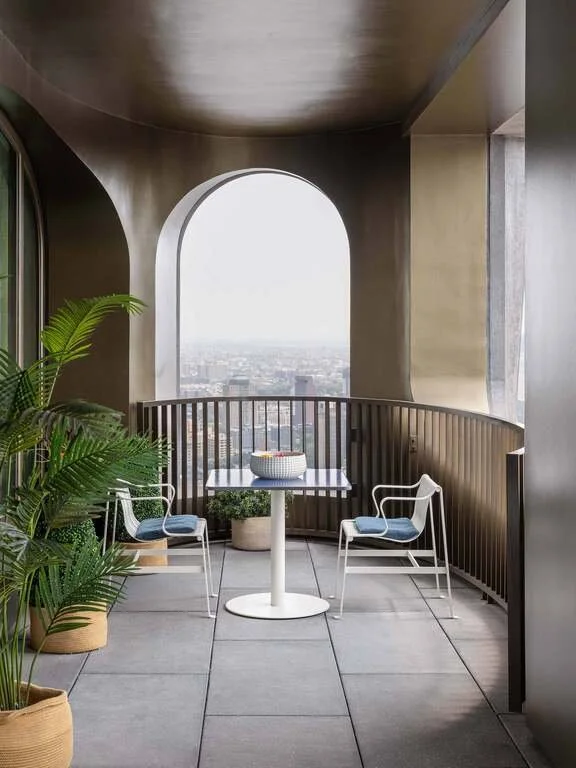 Balcony with white table, two white chairs with blue cushions, potted plant, and a city view through large arched window.
