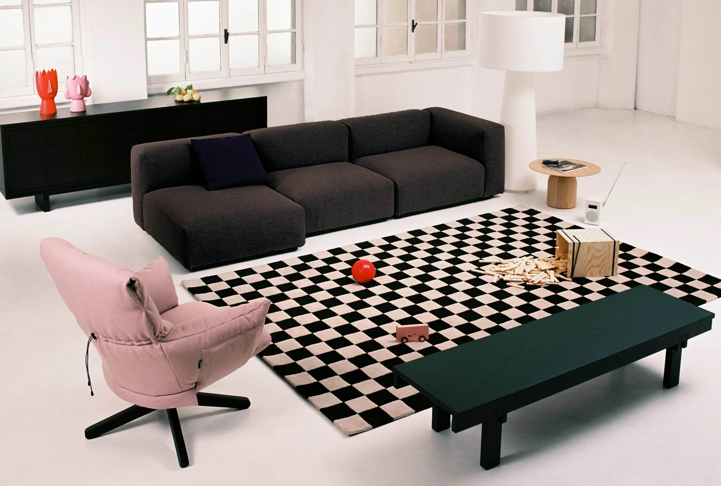 Modern living room with black sofa, pink armchair, black and white geometric rug, wooden side table, and decorative items on black console, large windows, and white walls.