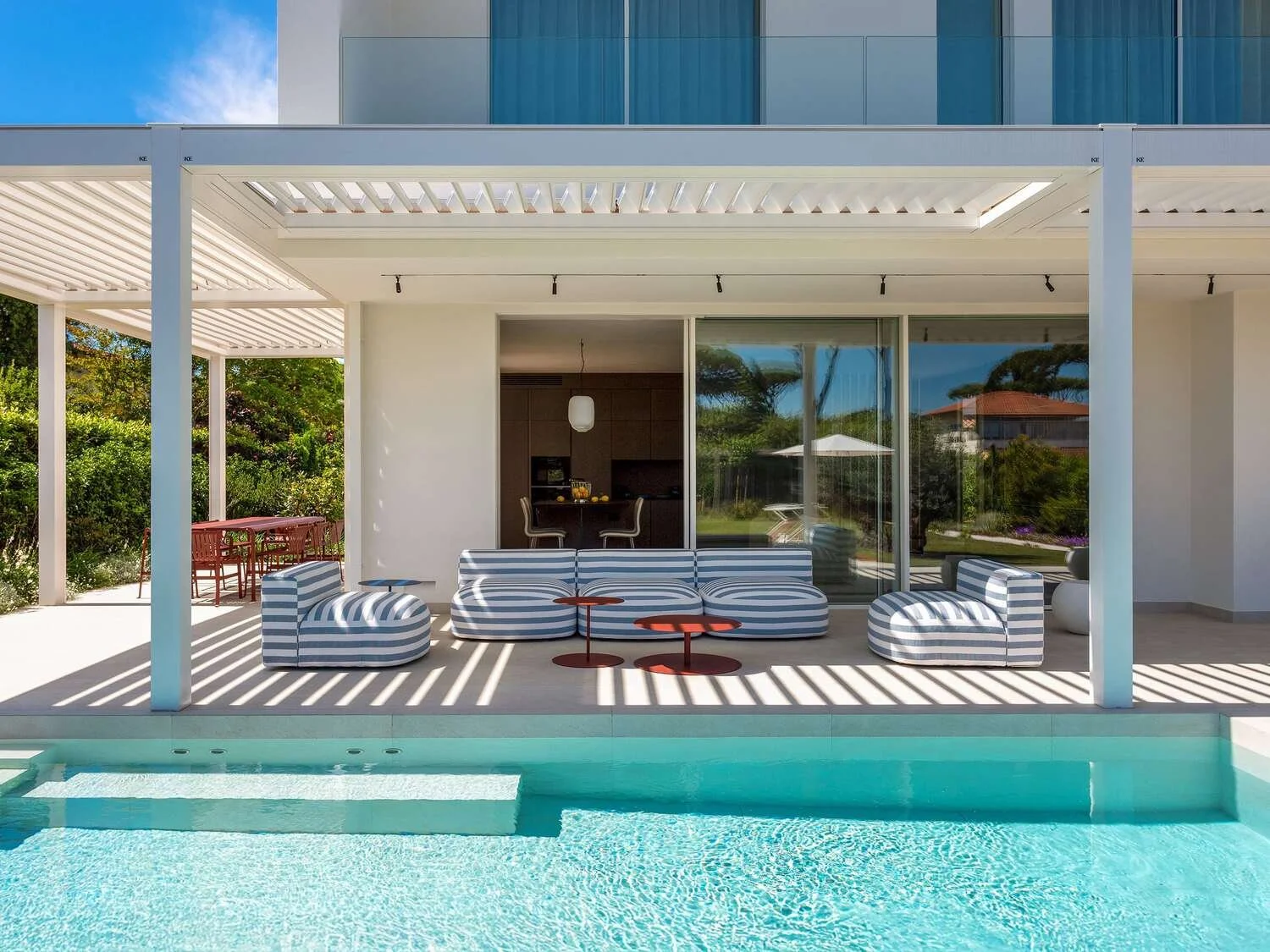 A modern outdoor patio with striped furniture, small round tables, and a pool in the foreground, with a house and lush greenery in the background.