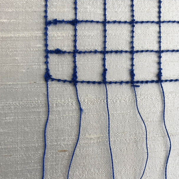 Close-up of blue embroidery thread on white fabric, forming a grid pattern with some loose threads hanging down.
