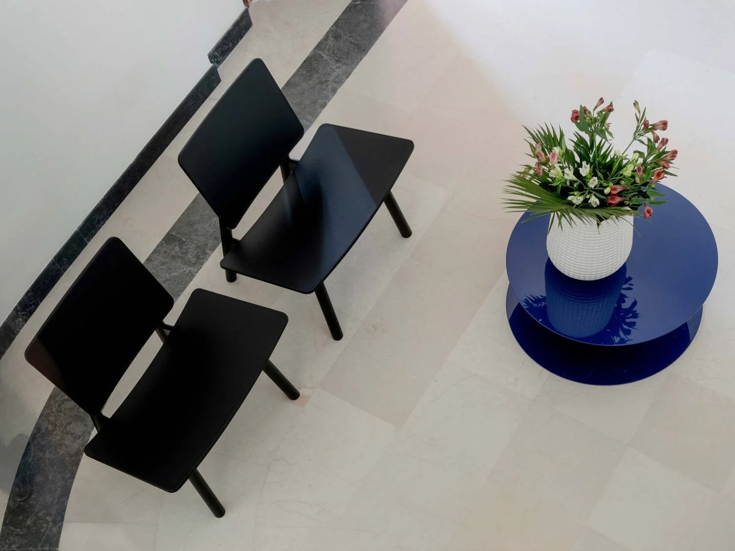 A minimalist interior scene with three black chairs aligned next to each other and a small round blue table with a white textured vase filled with pink and white flowers and green leaves. The floor is light-colored tile with a darker strip along the 
