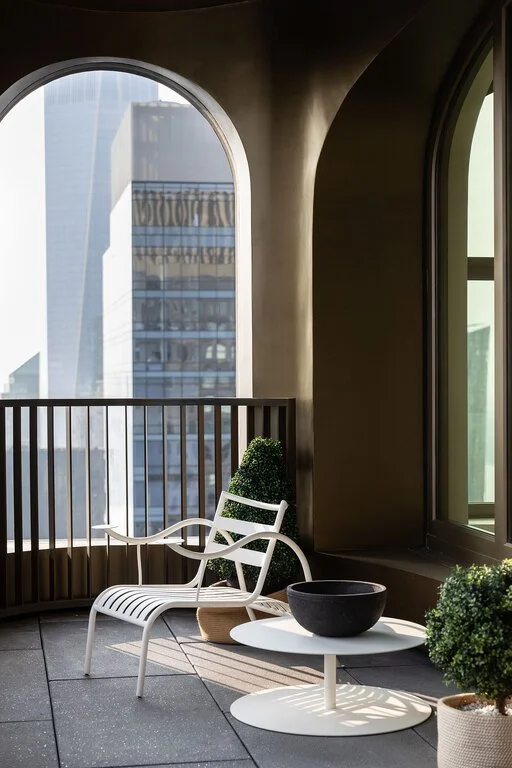 Balcony with modern white chair, black bowl, and potted plants, overlooking skyscrapers through large arched and rectangular windows.