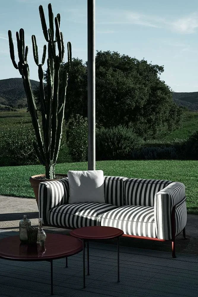A black and white striped sofa with a white pillow on it, located outdoors on a patio. Behind it is a large cactus in a terracotta pot and a scenic landscape with green hills and trees.