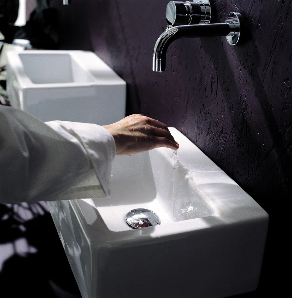 Person washing their hands under a running faucet in a white sink with a black textured wall behind.