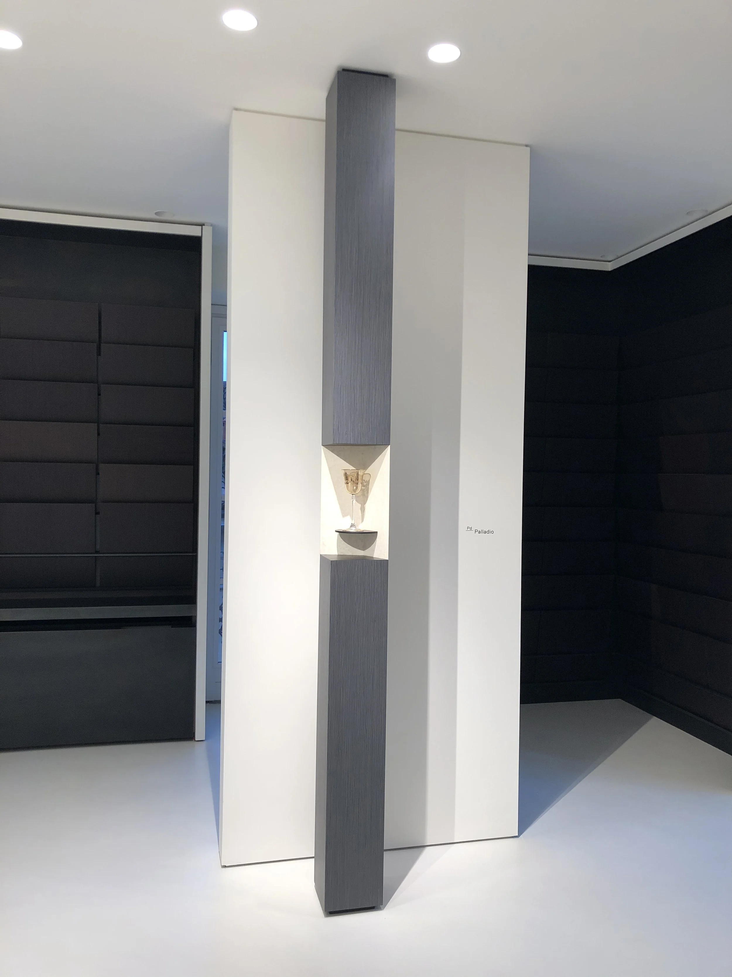 Modern interior space with black padded wall, white wall with a display shelf and a gray column with a small glass cup on top.