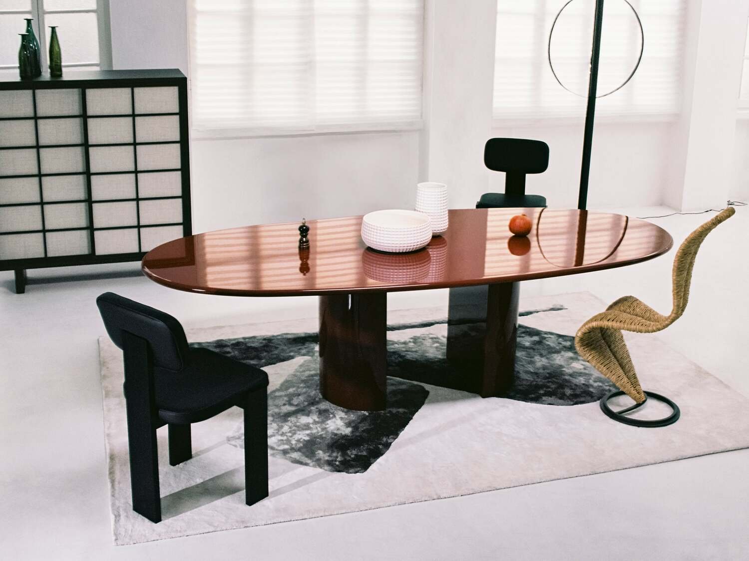 Modern dining room with an oval wooden table, black and woven chairs, decorative bowls, and a black sideboard with glass vases, under natural light and white window blinds.