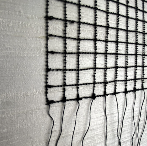 Close-up view of a black grid-like material with small square openings, attached to a textured white surface, with loose strings hanging below.