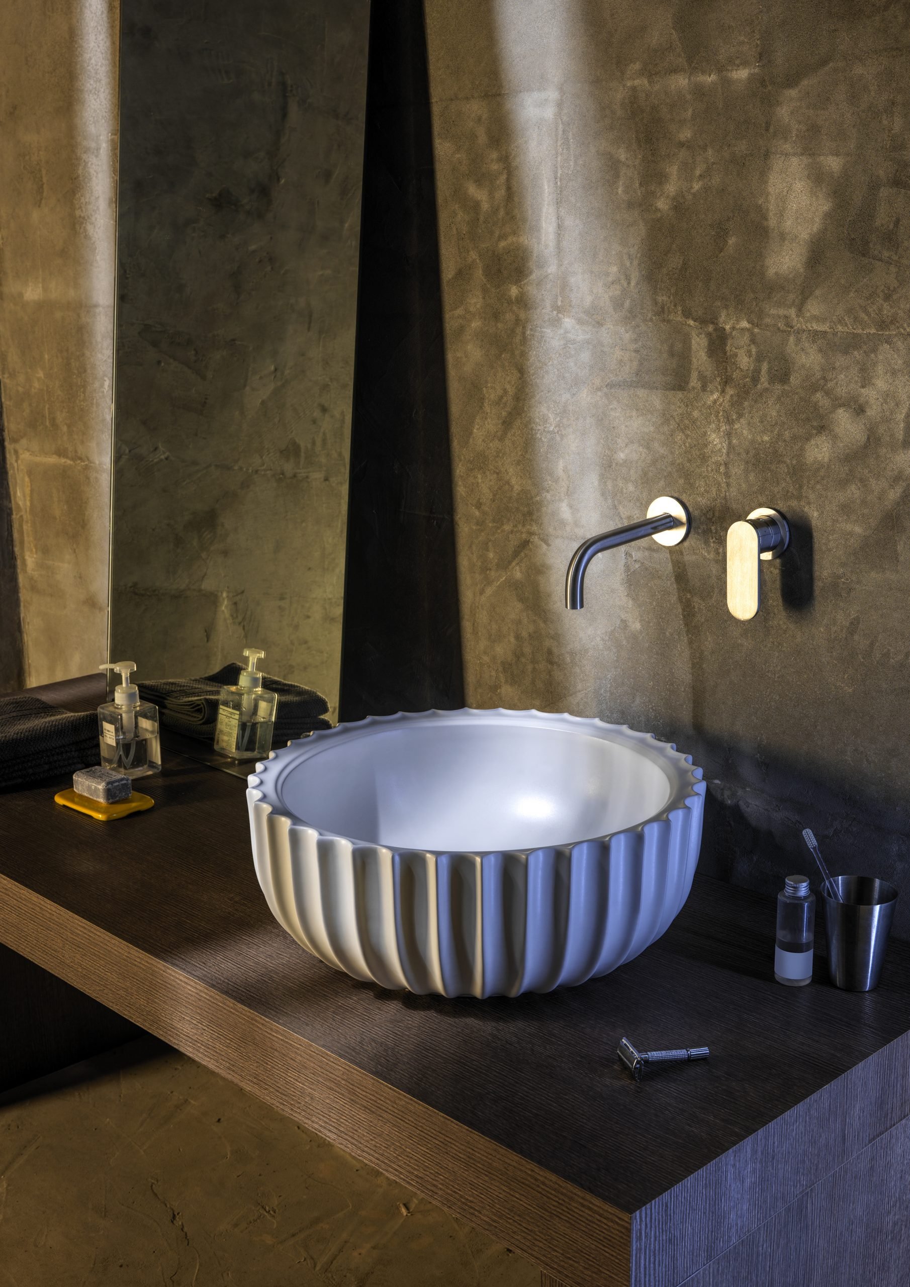 Modern bathroom vanity with a round, fluted white sink bowl, a wall-mounted faucet, and toiletries including soap, hand sanitizer, and a brush on a dark wood countertop. The background features a wall with gray textured tiles.