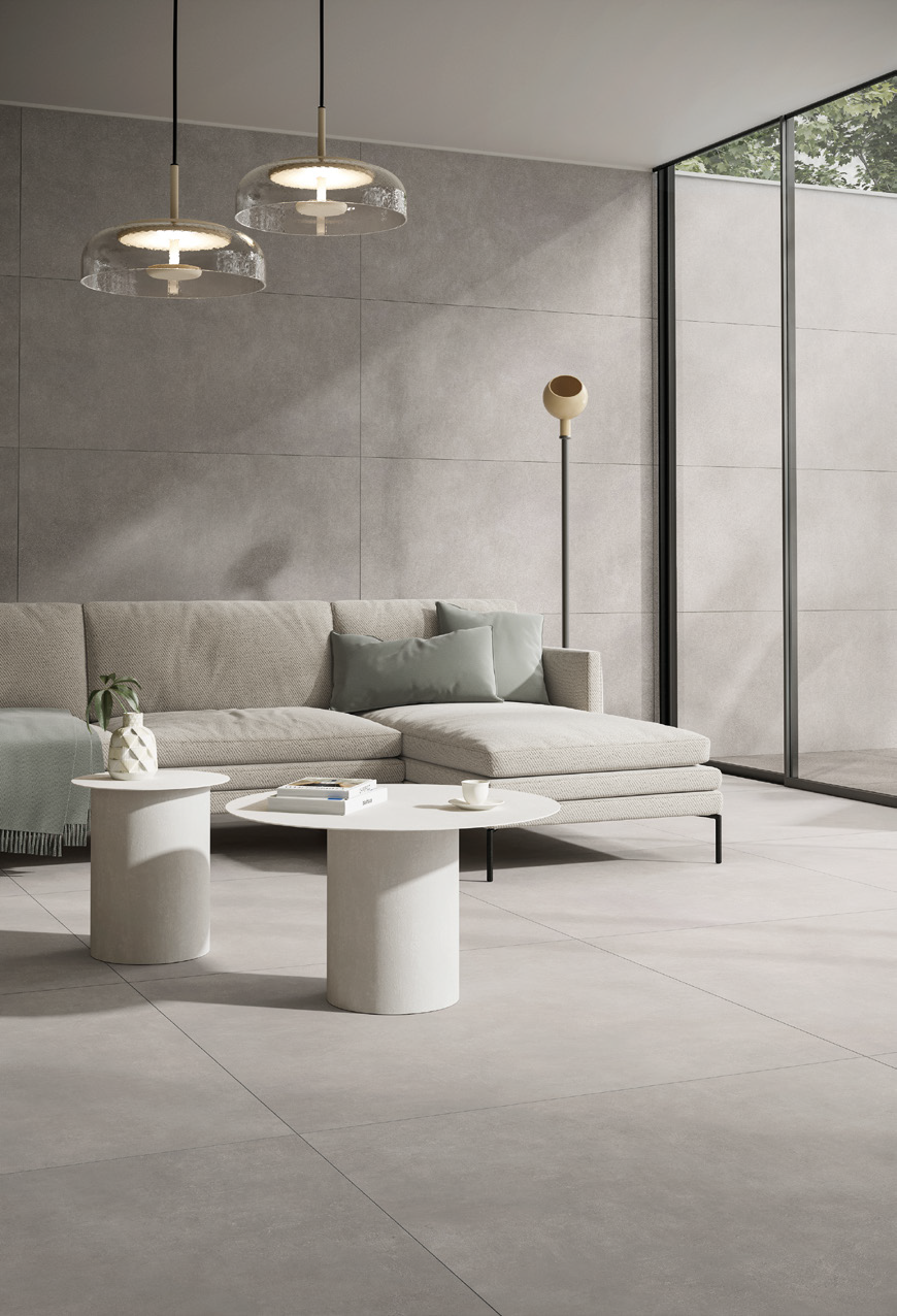 Minimalist living room with a light beige sofa, two modern pendant lights, a floor lamp, a small side table with a plant, and a round coffee table, all in neutral tones, with large glass windows letting in natural light.