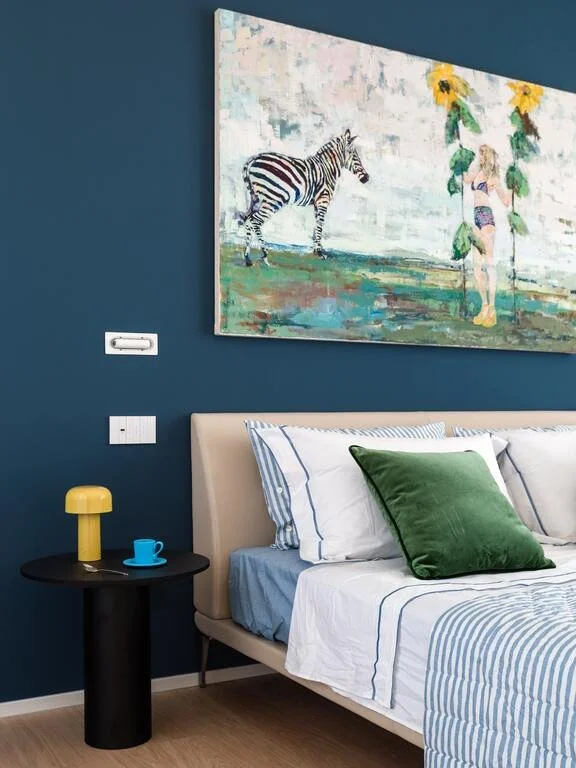 A bedroom with navy blue walls, a large colorful painting of a zebra and a person with sunflowers, a beige bed with striped white and blue bedding, a green pillow, and a black nightstand with a yellow lamp, a blue cup, and a yellow mushroom-shaped de