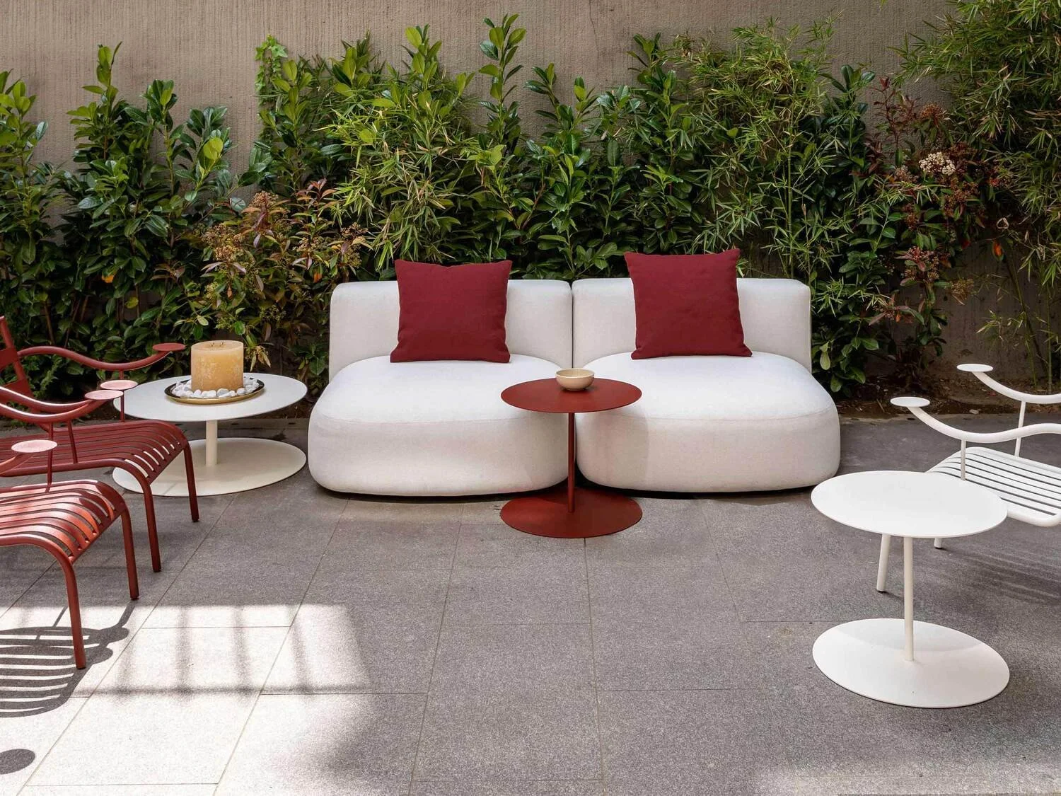 Outdoor seating area with a white curved sofa, two red throw pillows, a small round red table, and surrounding modern furniture including white and red side tables and chairs, with a lush green hedge background.
