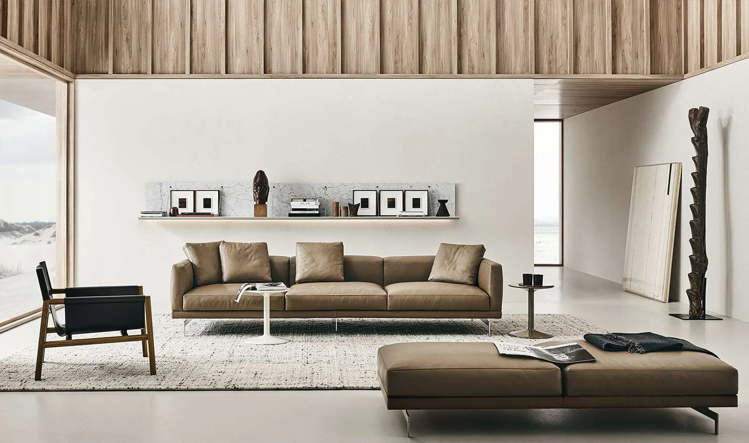 Modern living room with leather sofa, black chair, ottoman, and minimalist decor, featuring a large window with a view and abstract artwork.