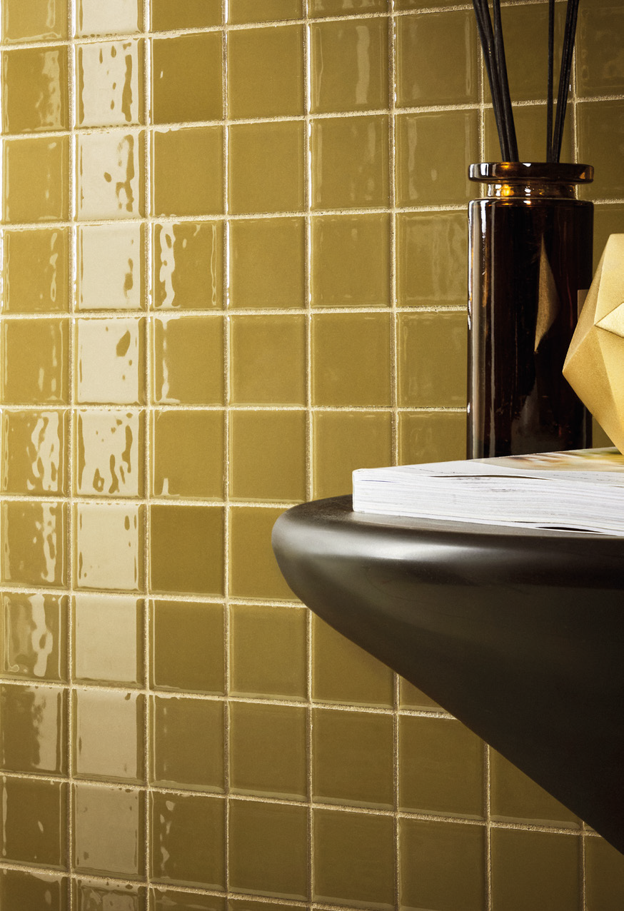 Close-up of a black countertop with a white book, a dark glass vase, and a yellow box against a yellow tiled wall.
