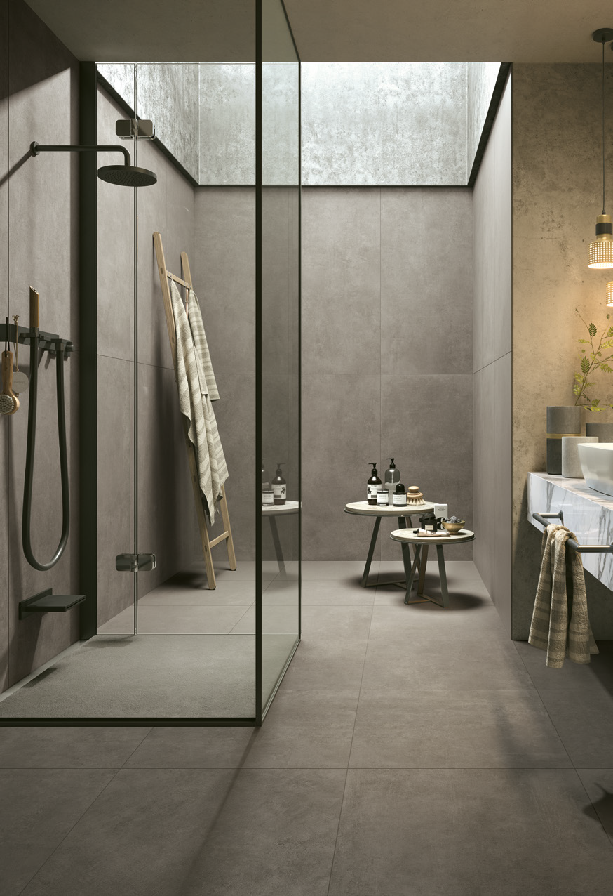 Modern minimalist bathroom with a glass-enclosed shower area, gray concrete walls, a high ceiling with a window, and a vanity with a sink. Decor includes small tables with toiletries and a towel hanging on the vanity.