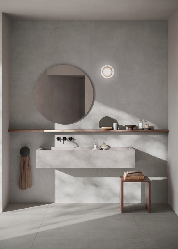 Minimalist bathroom with a concrete sink, round mirror, wall-mounted faucet, wooden shelf, and small wooden stool with folded towels.