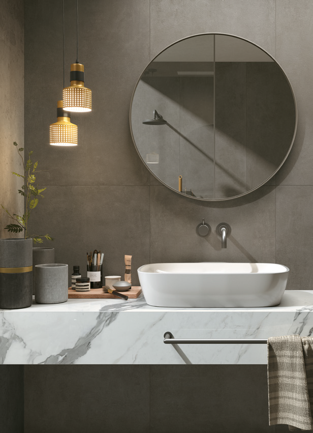Modern bathroom with a round mirror, wall-mounted showerhead, and white rectangular vessel sink on a marble countertop. Three hanging pendant lights, gray small planters with greenery, and toiletries on a wooden tray.