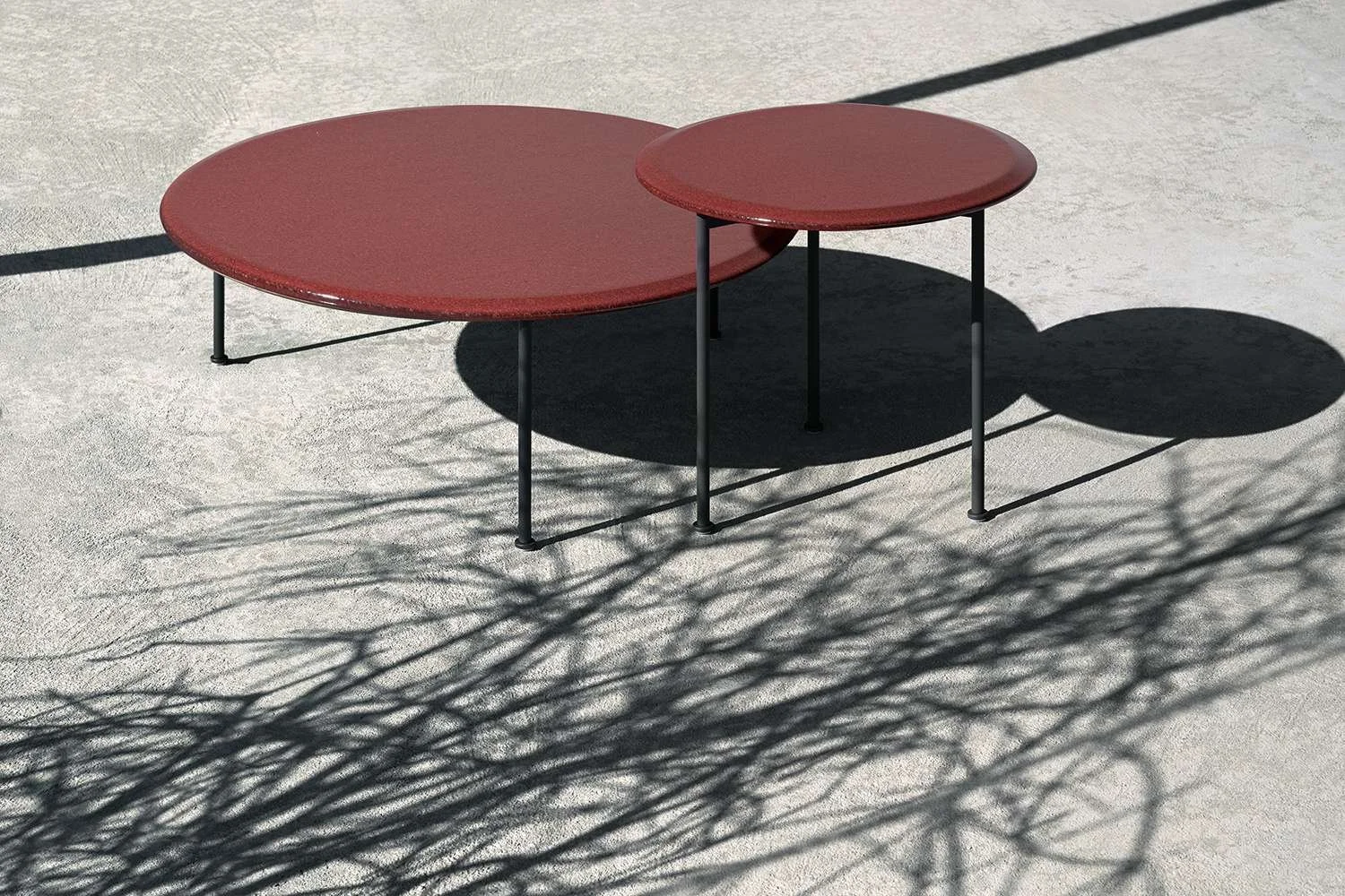 Two round, red tables with black metal legs on a concrete surface; shadows of leafless tree branches are cast across the ground and tables, creating a pattern.