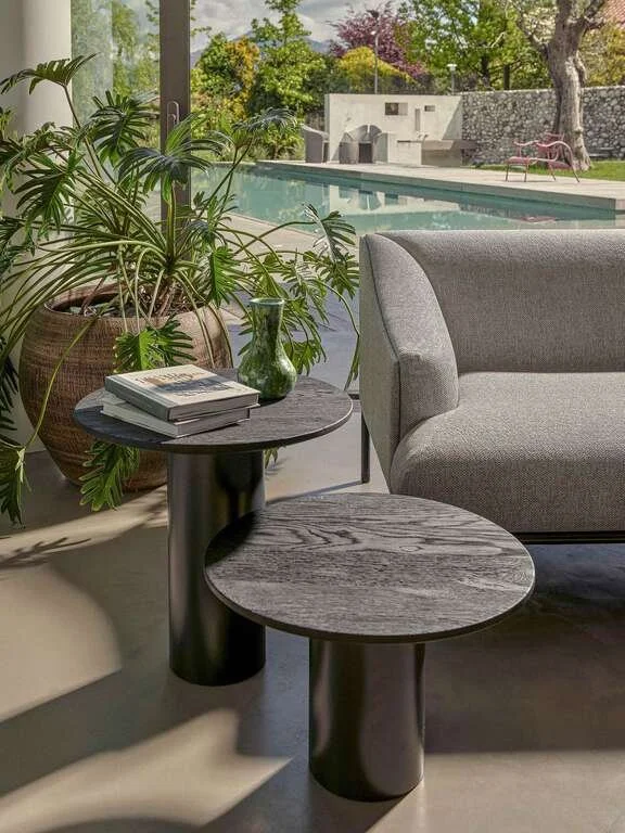A cozy indoor living space with a large potted plant, two nested round wooden tables with black bases, a stack of books, and a gray upholstered couch. Through the window, there is a backyard with a swimming pool, lush trees, and outdoor lounge chairs
